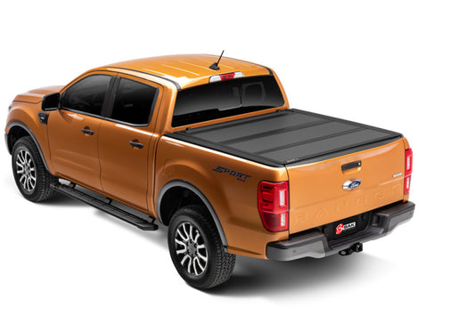 An orange pickup truck with a covered bed is shown from a rear three-quarter angle. The truck features black accents, a "Sport" decal on the right side, and showcases a sleek, heavy-duty BAKFlip MX4 Matte Finish aluminum tonneau cover with UV resistance designed for the 19-20 Ford Ranger 5ft Bed by BAK. The vehicle has large, rugged tires and a rear-mounted hitch.