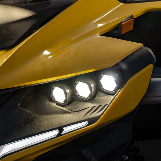 Close-up of a yellow vehicle's front lights, featuring the Baja Designs 2024 Can-Am Maverick R Triple S1 Unlimited Headlight Kit, incorporating three hexagonal LED lights. The sleek design showcases angular lines with a black and yellow color scheme, capturing the sporty aesthetic of a Can-Am Maverick.