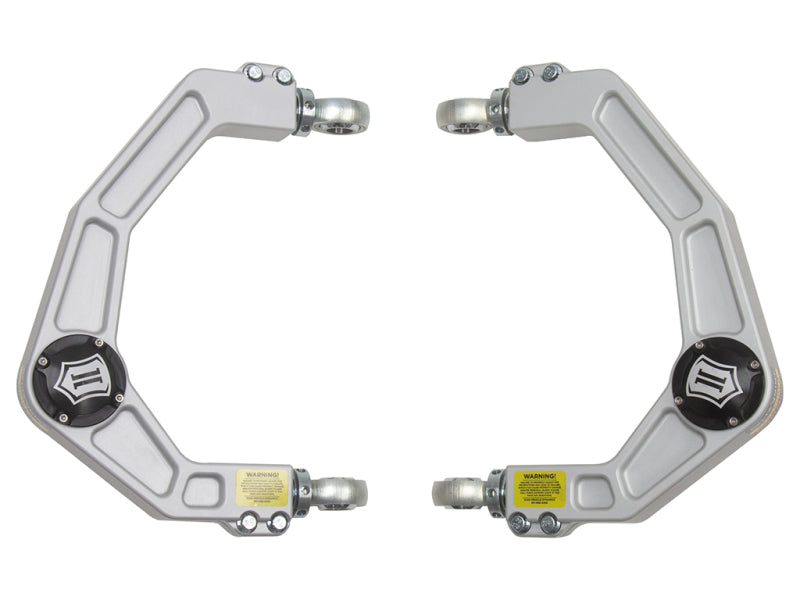 Image of two ICON 2021+ Ford F-150 Billet Upper Control Arms with bolts and labeled warning stickers. The symmetrical upper control arms are positioned facing each other, featuring a geometric design and a circular component in the center. These ICON control arms boast corrosion resistance for enhanced durability.