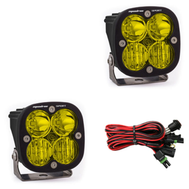 Two amber square LED light pods from Baja Designs' Squadron Sport series are displayed, featuring black casings and metal brackets. Below them is a wiring harness with black connectors and red and black wires, highlighting the durable design that the brand is known for.