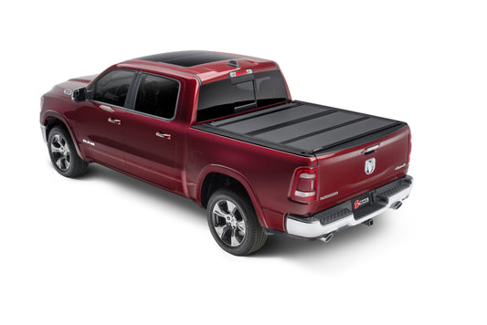 A red crew cab pickup truck is visible from the rear left side, highlighting its sleek design and the BAK 19-20 Dodge Ram 1500 (New Body Style w/o Ram Box) 6ft 4in Bed BAKFlip MX4 Matte Finish black tonneau cover over the truck bed. The truck features shiny alloy wheels, a black roof, and chrome accents on its trim, grille, and side mirrors.