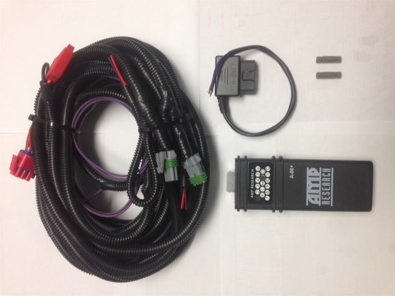 A black coiled wiring harness with various colorful connectors, a small grey rectangular component with wires, two small cylindrical items, and a black rectangular device labeled "AMP Research PowerStep" are arranged on a white background, showcasing parts from the AMP Research 2009-2014 Ford F150 PowerStep Plug N Play Conversion Kit by AMP Research.