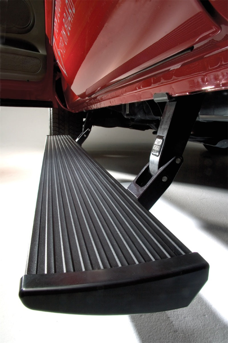 A close-up view of an AMP Research 2016-2017 Nissan Titan / Titan XD PowerStep Plug N Play running board under the side of a red truck, providing a step for easier access into the vehicle. The black running board has a ridged, non-slip surface and is supported by metal brackets. It comes with a 0-mile warranty.