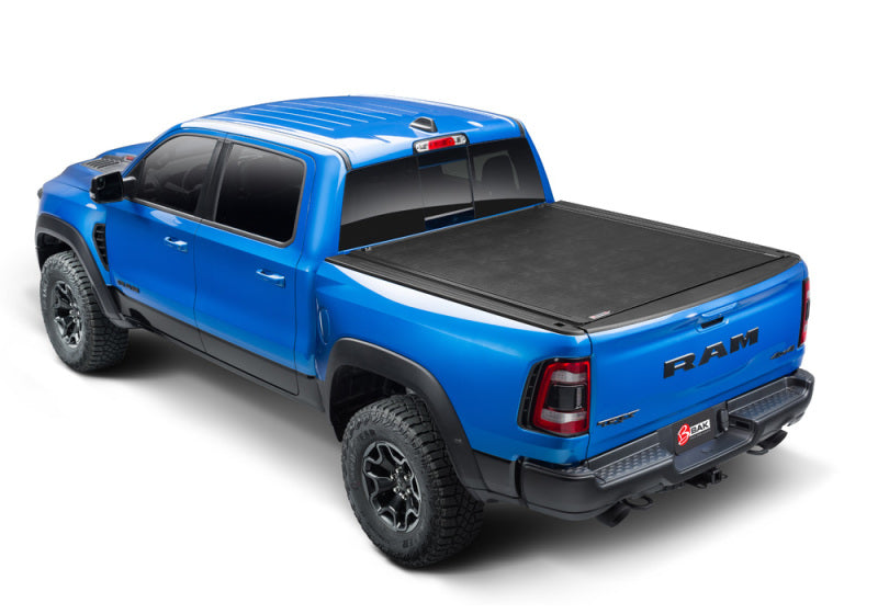 A bright blue Dodge Ram 1500 pickup truck from the 09-18 series, equipped with a BAK Revolver X2 aluminum roll-up cover on its 5ft 7in bed, is shown from a high rear three-quarter angle. The truck features a crew cab, rugged tires, and a tailgate adorned with the Ram logo. Distinctive rear lights and a visible exhaust pipe enhance its robust appearance.