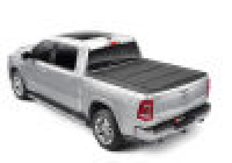 A silver pickup truck with a black BAK 09-18 Dodge Ram 1500 (19-20 Classic Only) 5ft 7in Bed (w/ Ram Box) BAKFlip MX4 Matte Finish tonneau cover over the bed, viewed from the rear-left side. The vehicle has four doors and tinted windows, featuring a heavy-duty aluminum truck bed cover for added durability.