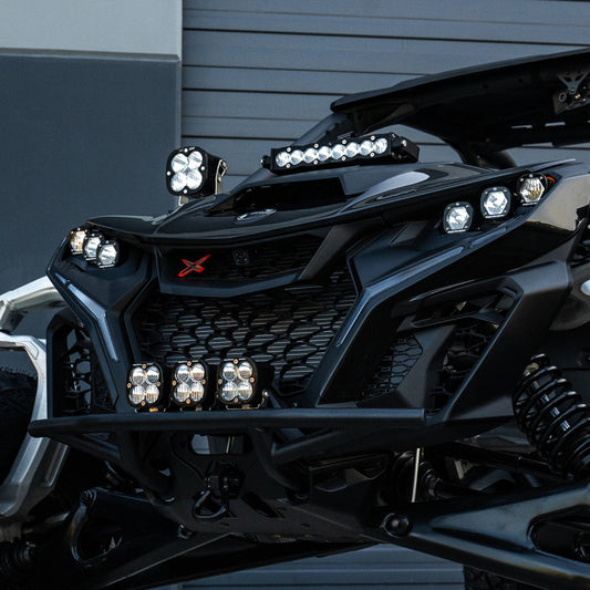 Close-up of a Baja Designs Can-Am Maverick R Clear 10in S8 Shock Tower Kit showcasing its rugged off-road front with multiple LED lights. The vehicle features an aggressive grille design, black and silver detailing, and a red "X" emblem. In the background, a gray garage door is visible.