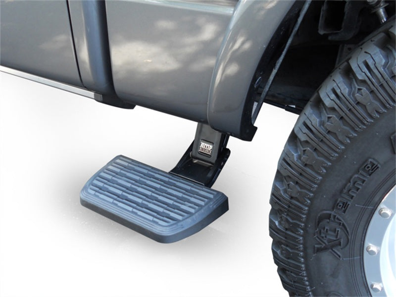 A close-up view of the AMP Research 19-22 Chevrolet/GMC Silverado/Sierra 1500 Short Bed BedStep2 - Black, mounted beneath the truck's body. The step is extended, showcasing its textured tread surface for grip, ensuring easy toolbox access. Part of a large off-road tire with rugged tread is visible in the foreground on the right.