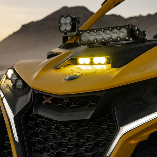 Close-up of a yellow off-road vehicle with a Baja Designs 2024+ Can-Am Maverick R S2 Pro Hood Light Kit. The rugged design highlights a prominent grill and headlights, standing against a mountainous backdrop at dusk, enhancing its adventurous appeal.