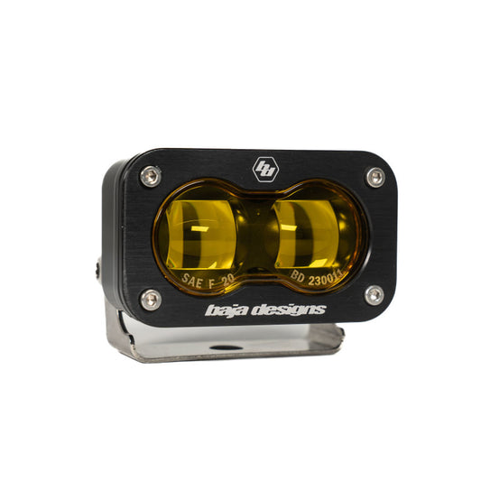 The Baja Designs 19-22 Dodge RAM 2500/3500 S2 SAE Fog Pocket Light Kit - Amber features black and yellow lights with two round amber lenses. Mounted on a silver metal bracket, it has a black faceplate adorned with the Baja Designs logo, screws at each corner, and model information. This kit is perfect for enhancing your Dodge RAM 2500/3500's visibility in foggy conditions.