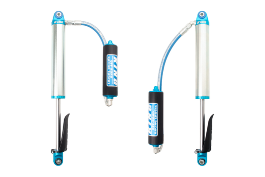 Image showing a pair of King Shocks 2019+ Chevrolet Silverado 1500 (Trail Boss) Rear 2.5 Dia Remote Reservoir Shocks. The shocks are positioned side by side, showcasing their silver cylindrical bodies with blue accents and black hoses connecting to the reservoirs labeled “King Shocks.” These bolt-on suspension components promise superior wheel control and performance for your vehicle.