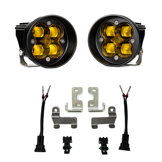 The image showcases the Baja Designs 12-21 Toyota Tacoma/Tundra/4Runner Squadron-R Fog Pocket Light Kit - SAE Amber, featuring a pair of round LED fog lights with yellow SAE Amber lenses, viewed from the front. Below these lights are mounting brackets, wiring connectors, and screws required for installation. This kit by Baja Designs is specifically designed to enhance visibility in foggy or low-light conditions.