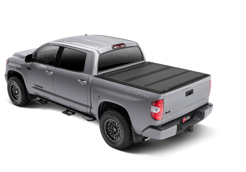 A silver Toyota Tundra featuring a black BAK 2022+ Toyota Tundra 6.5ft Bed BAKFlip MX4 Bed Cover shown from a rear three-quarter view. The truck includes black alloy wheels, running boards, and tinted windows, with "TRD PRO" text visible on the truck bed. The background is plain white.