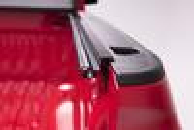 The image shows a partial view of a red pickup truck bed featuring the BAK 19-20 Chevy Silverado 5ft 8in Bed (New Body Style) BAKFlip F1 hard folding truck bed cover by BAK. The cover is partially retracted, revealing the ribbed surface of the truck bed. The truck is positioned in a well-lit environment, showcasing the sleek Fiberglass Reinforced Polymer design.