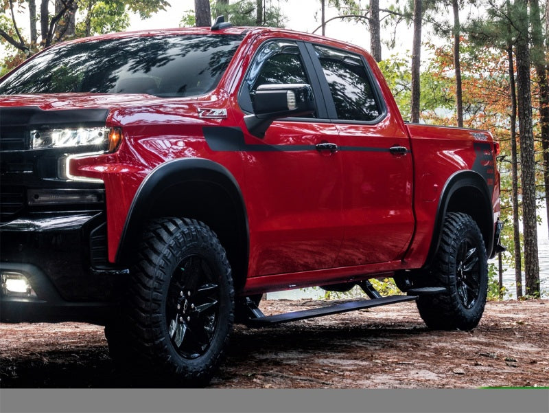 An AMP Research 19-21 Chevy Silverado 1500 Crew Cab PowerStep Plug N Play - Black (w/OEM Illumination) parked in a wooded area. The double cab pickup has black wheels with rugged tires. The background shows tall trees and a glimpse of a lake, conveying a sense of adventure and outdoor exploration.