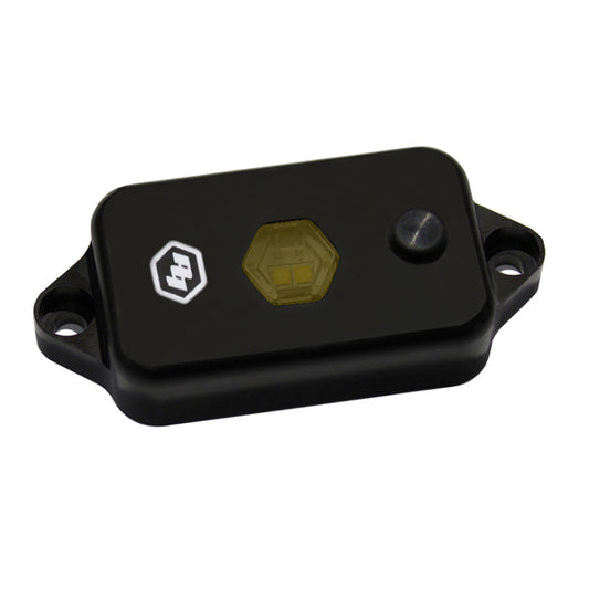 A black rectangular electronic sensor device features a hexagonal light indicator similar to the Baja Designs LED Dome Light w/Switch - Amber, with a small button on its surface. It includes mounting holes on either side for easy installation and emits an amber glow reminiscent of those from Baja Designs.