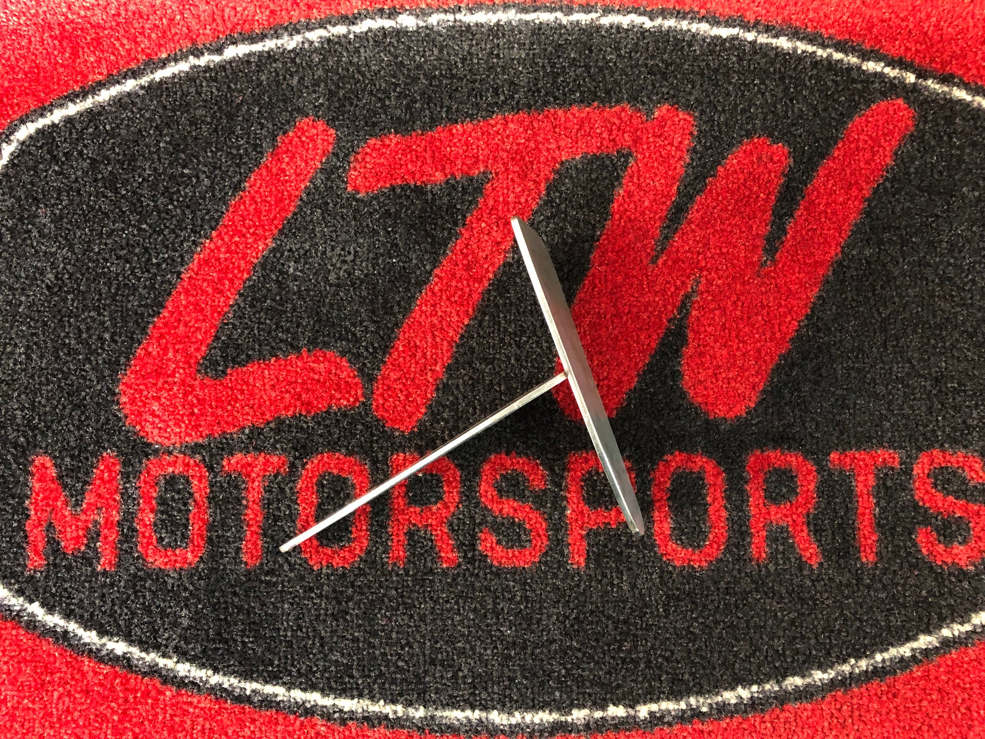 A metal valve rests horizontally across a red and black mat with "LTW Motorsports" prominently displayed in bold red letters on a black oval background, next to LTWBMC1 body mount chop plates for a Toyota Tacoma.