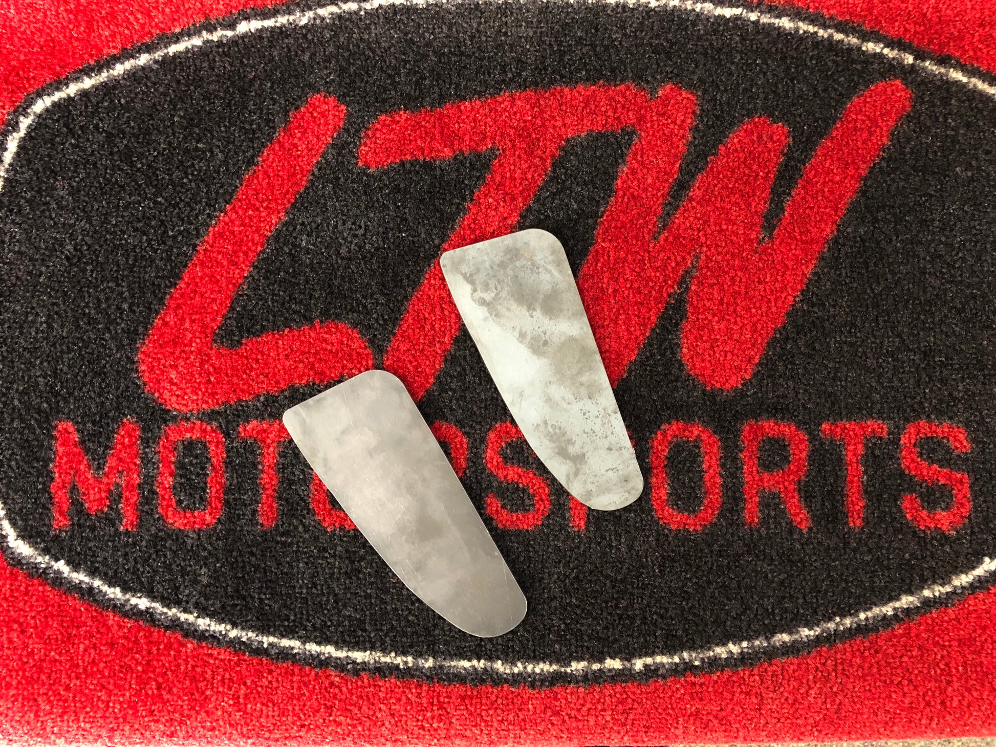 A metal valve rests horizontally across a red and black mat with "LTW Motorsports" prominently displayed in bold red letters on a black oval background, next to LTWBMC1 body mount chop plates for a Toyota Tacoma.