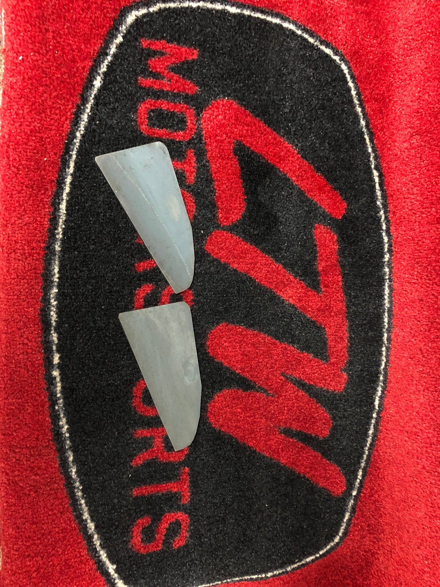 Two small, thin, gray metal or plastic pieces, likely body mount chop plates for a Toyota Tundra with oversize tires, rest on a red and black cloth bearing the "LTW Motorsports" logo. The cloth features a black oval background around the logo. The pieces are similar in shape and size. These parts are labeled as model LTWBMC2.