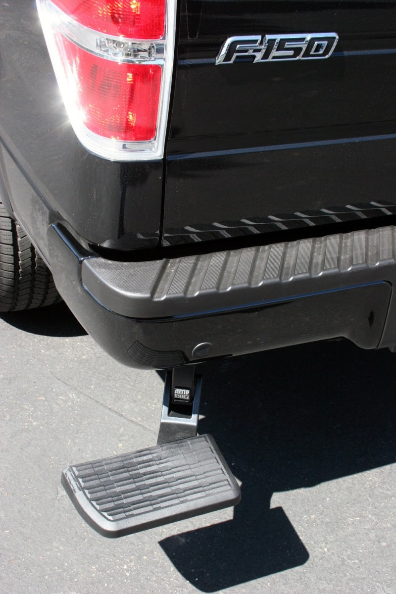 A black AMP Research 2006-2014 Ford F150 BedStep extends from the rear bumper of the truck, providing easier access to the truck bed. The retractable step lowers downward, resting just above the ground while the truck is parked on a paved surface.