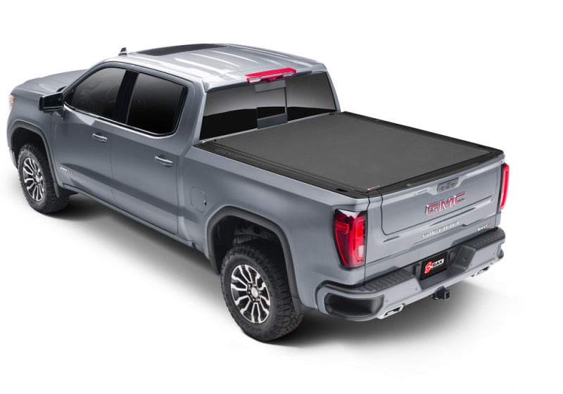 A sleek silver GMC pickup truck is shown from a rear-side angle. The truck is equipped with a premium BAK 2023+ Chevy Colorado Revolver X4s 5.2ft Bed Cover over the bed. The vehicle has tinted windows, stylish alloy wheels, and visible red tail lights. The background is white and plain.