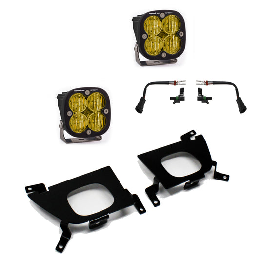 The Baja Designs 2019 Silverado 1500 Pro Fog Pocket Mount Kit includes a pair of yellow square LED fog lights, two black metal mounting brackets, and a set of wiring harnesses with connectors. This off-road lighting kit is specifically designed for the Silverado 1500, with components arranged separately on a white background.