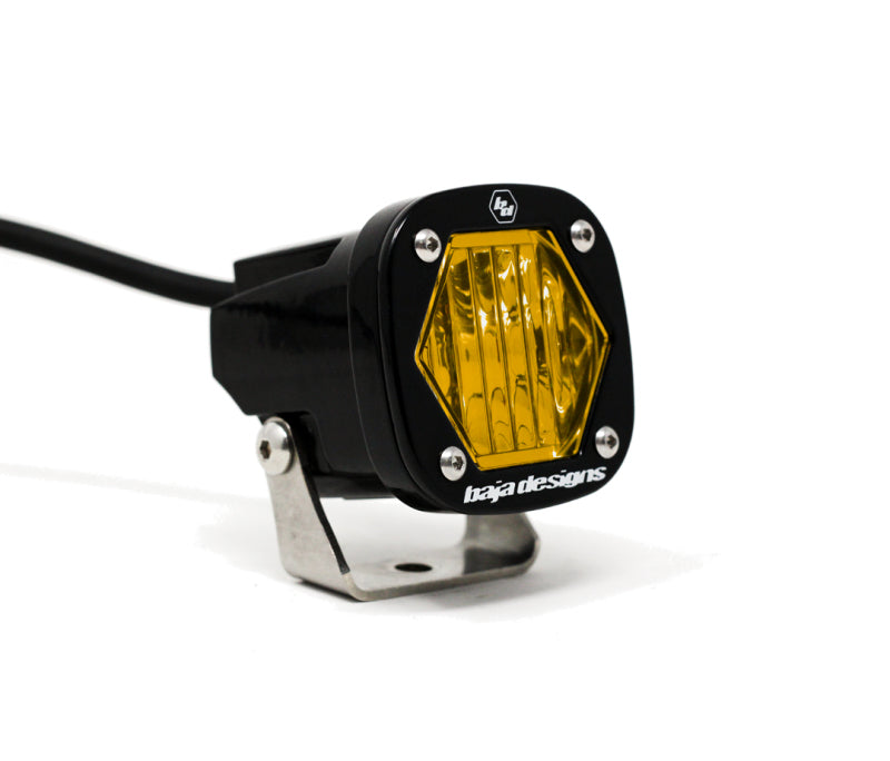 A close-up of the Baja Designs S1 Amber Wide Cornering LED Light with a hexagonal lens, mounted on a bracket. The "Baja Designs" label is visible on the front, and a cable extends from the back.