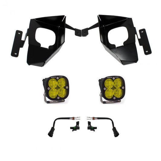 A set of black mounting brackets with cut-out holes, two square-shaped amber LED fog lights with four LED bulbs each and black casings, and two wiring harnesses from the Baja Designs 16-18 Silverado Squadron Sport Fog Pocket Kit W/C - Amber are laid out against a white background.