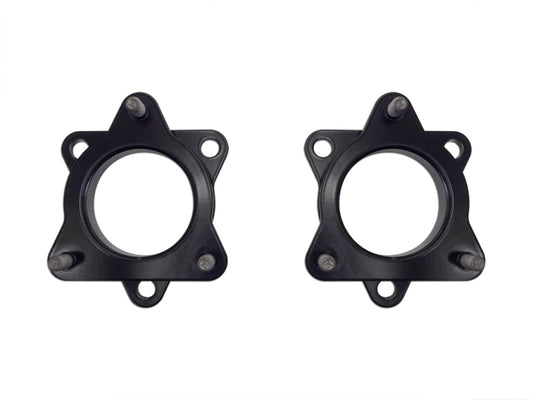 Two black metal automotive parts with a circular center and a star-like, 5-point outline are displayed against a white background. Each has threaded bolts on the points and circular holes on two sides. These items, from the ICON 2004+ Ford F-150 2in Spacer Kit by ICON, are compatible with the Ford F150 IVD Leveling Spacer Kit for a 2-inch lift and are placed side by side, facing forward.