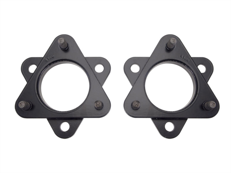 Two identical black metal plates, each with a circular center hole and three protruding threaded bolts arranged in a triangular pattern. These ICON 2005+ Toyota Tacoma / 2007+ Toyota FJ / 2002+ Toyota 4Runner 2in Spacer Kits also feature three smaller holes near the bolts for additional mounting options.